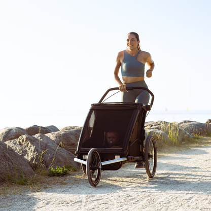 Thule Kinderanhänger beim Joggen – sportliche Person joggt mit schwarzen Thule Chariot Cross mit großem Vorderrad-Set auf sonniger Straß