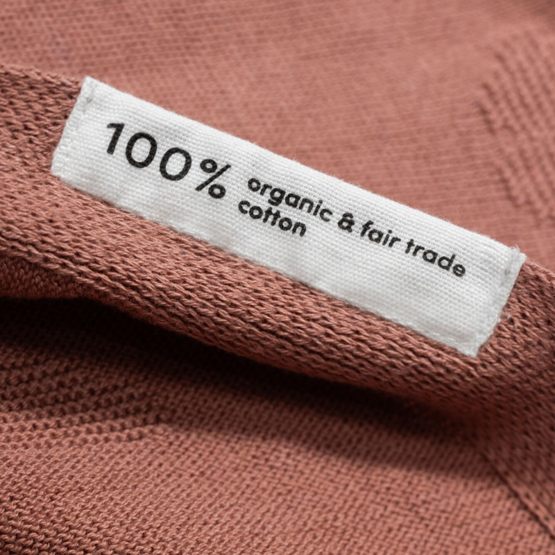 Nahaufnahme eines Stoffetiketts mit der Aufschrift ‚100% organic &amp; fair trade cotton‘ auf einem terrakottafarbenen Strickstoff.